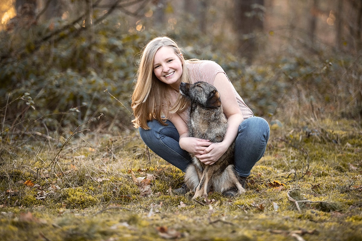 lachende blonde Frau hockt auf dem Waldboden und hält ihren Hund im Arm
