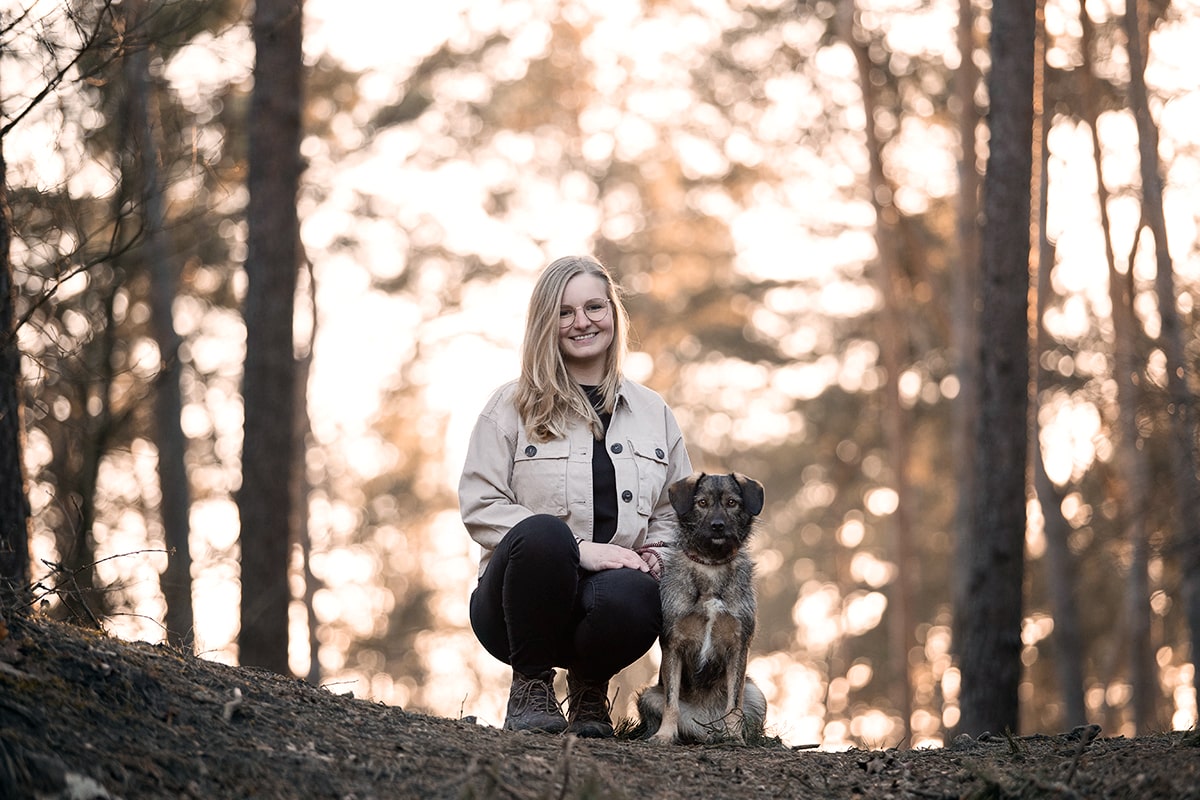 Blonde Frau mit Brille und ihr Mischlingshund im Wald bei Sonnenuntergang